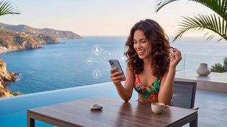 Kostume Rio de Janeiro USA - A woman with long wavy brown hair sits at an outdoor table by the ocean, smiling while holding a smartphone. She is wearing a colorful floral bikini top.
