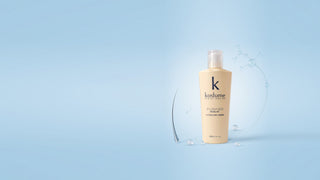 A bottle of Kostume hair mask  by Kostume RIo de Janeiro in a pale beige shade, standing on a light blue reflective surface with a subtle water splash graphic behind it.